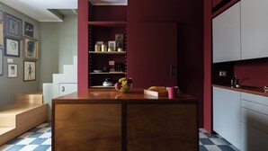 Une cuisine avec un îlot central en bois, un panier de fruits et une boîte à pain. Des tableaux encadrés ornent le mur, et un escalier mène à un autre niveau.