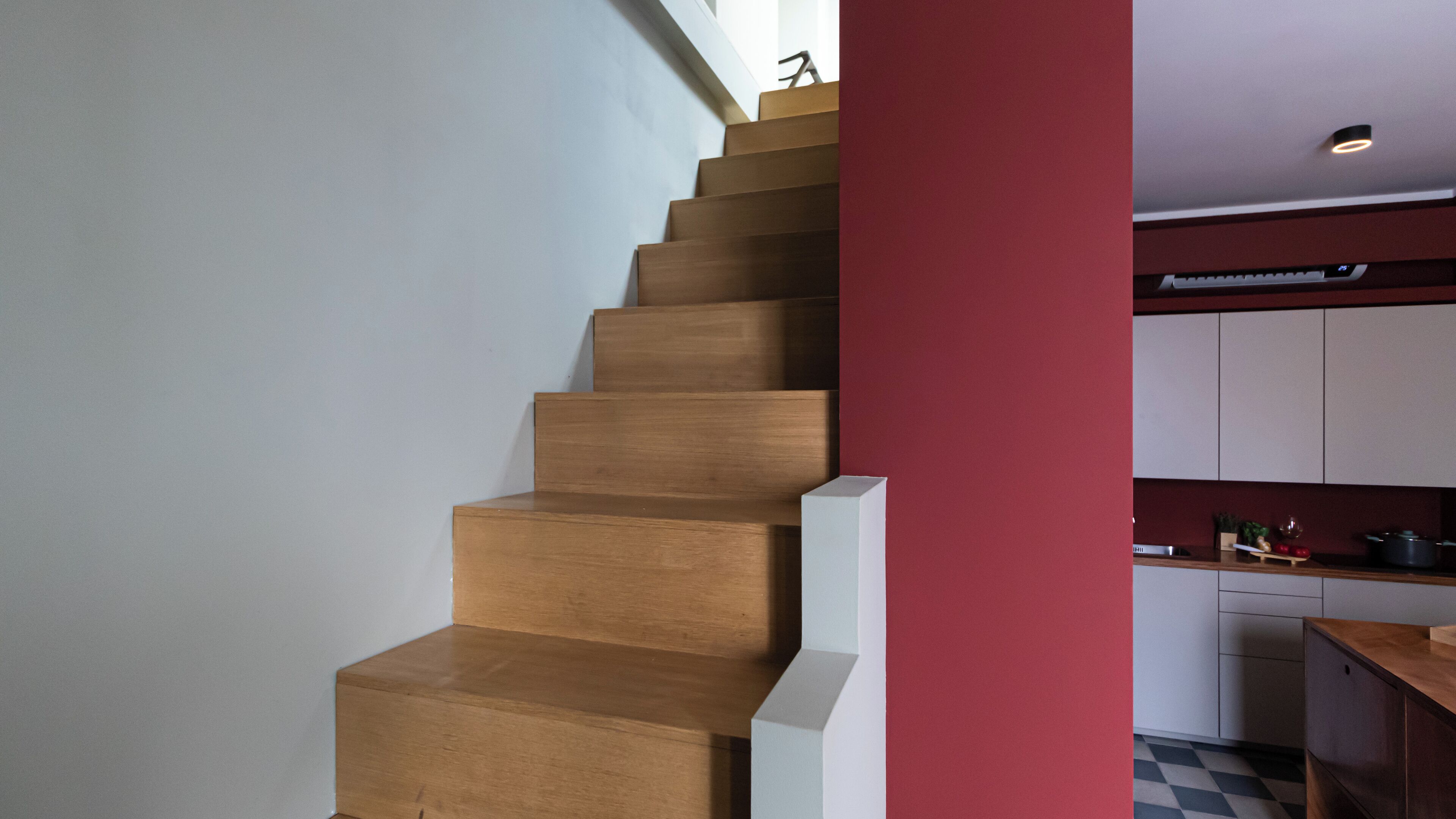 Una escalera de madera que lleva a una zona de cocina con armarios blancos y una pared roja decorativa.