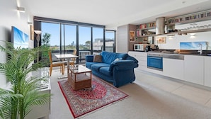 A modern apartment with a dining area, a blue sofa, a kitchen with white cabinets, and a large window offering a city view. - Eclectic 1-bed Apartment With Pool and Gym (Melbourne)