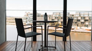 Varanda com mesa e duas cadeiras, com vista para um corpo de água e edifícios.