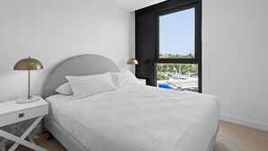 A modern bedroom with a large bed, a nightstand with a lamp, and a window offering a city view. - Executive 2-bed Apartment With Views, Gym and Spa (Melbourne)