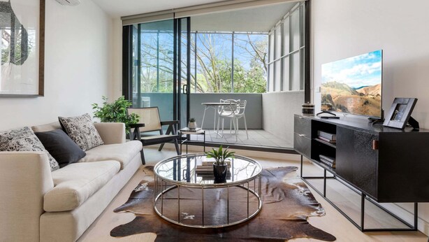 Ein modernes Wohnzimmer mit einer Couch, einem Glastisch, einem Lederteppich, einem Fernseher und einem Balkon mit Blick auf Bäume.