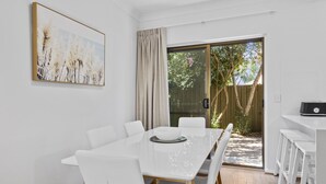A dining area with a white table and chairs, a painting on the wall, and a sliding glass door leading to an outdoor area. - Coastal 3-bed House With BBQ and Private Courtyard (Adelaide)