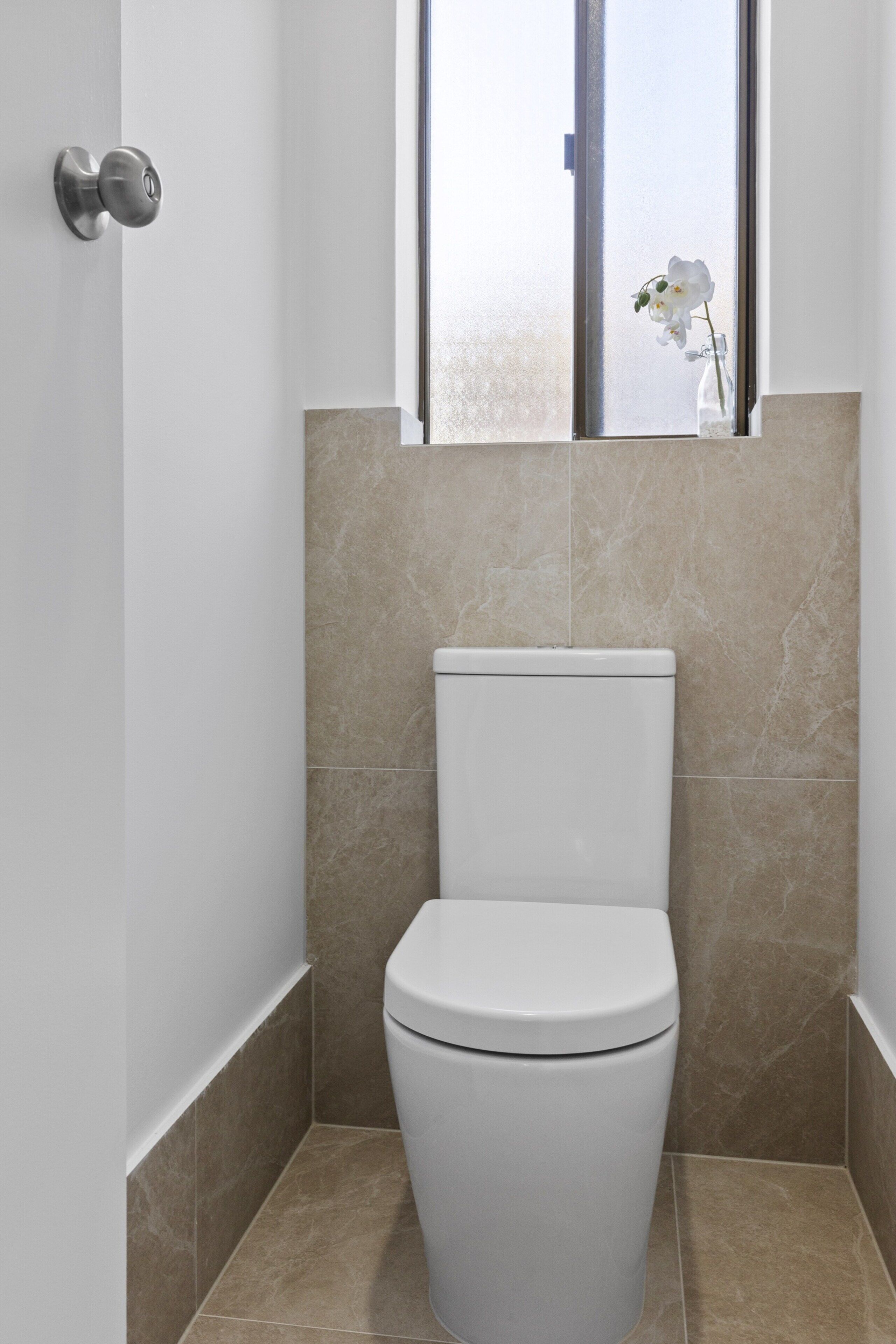 A modern bathroom with a white toilet, beige tiles, and a window with a view.