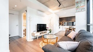 A modern living room with a grey sofa, a glass coffee table, and a flat-screen TV. There is a kitchen in the background with wooden cabinets and a dining area. - Stylish Southbank 1-bedroom With Pool and Gym (Melbourne)