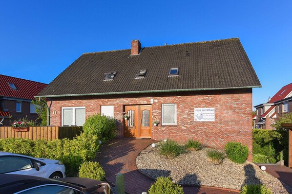 A two-story brick building with a sloped roof, a wooden door, and a small garden area with gravel and plants. - Ferienwohnung Balkon zur Sonnenseite in Bensersiel (Esens)