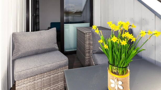 A balcony with a grey sofa, a wicker table, and a vase of yellow flowers. - Ferienwohnung Balkon zur Sonnenseite in Bensersiel (Esens)