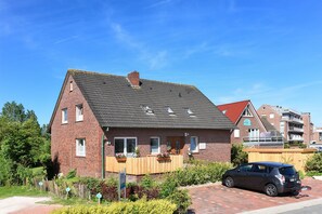A two-story brick house with a sloped roof, a wooden fence, and a car parked in the driveway. - Ferienwohnung Balkon zur Sonnenseite in Bensersiel (Esens)