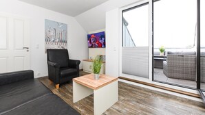 A modern living room with a black sofa, a wooden coffee table, and a black armchair. There is a painting on the wall, a television, and a view of the outdoors through a sliding glass door. - Ferienwohnung Balkon zur Sonnenseite in Bensersiel (Esens)