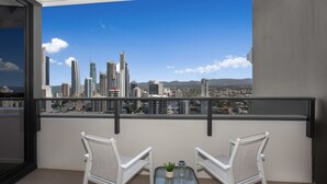 Un balcon avec deux chaises blanches et une petite table, d’où l’on peut admirer un paysage urbain avec de hauts immeubles et un ciel bleu dégagé.