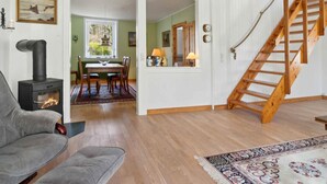 A cozy living room with a fireplace, a sofa, a dining area with a table and chairs, and a wooden staircase. - 5 Star Holiday Home in Allinge (Allinge)