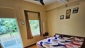 A bedroom with a bed, a ceiling fan, a window with curtains, a small table, and a door leading to a garden. - Prabhukrupa Tourist Farm (Alibag)