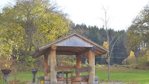 Un belvédère rustique en bois, doté d’un toit en pente, entouré d’un champ herbeux et d’arbres.