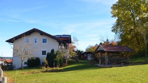 Une maison à deux étages avec un toit en pente, une petite dépendance attenante avec un toit rouge, et une cour verdoyante avec des arbres et des arbustes.