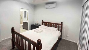 Un dormitorio con una cama de madera, un ventilador de techo, un equipo de aire acondicionado, una mesita de noche y un baño visible a través de una puerta abierta.