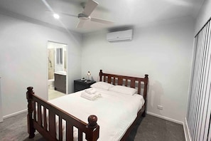 A bedroom with a wooden bed, a ceiling fan, an air conditioning unit, a nightstand, and a bathroom visible through an open door.