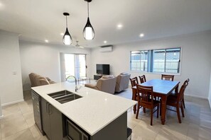 A modern kitchen with a central island, dining area with wooden chairs, a flat-screen TV, and a large window. - New  4Bed House master with ensuite and A/C (Collingwood Park)