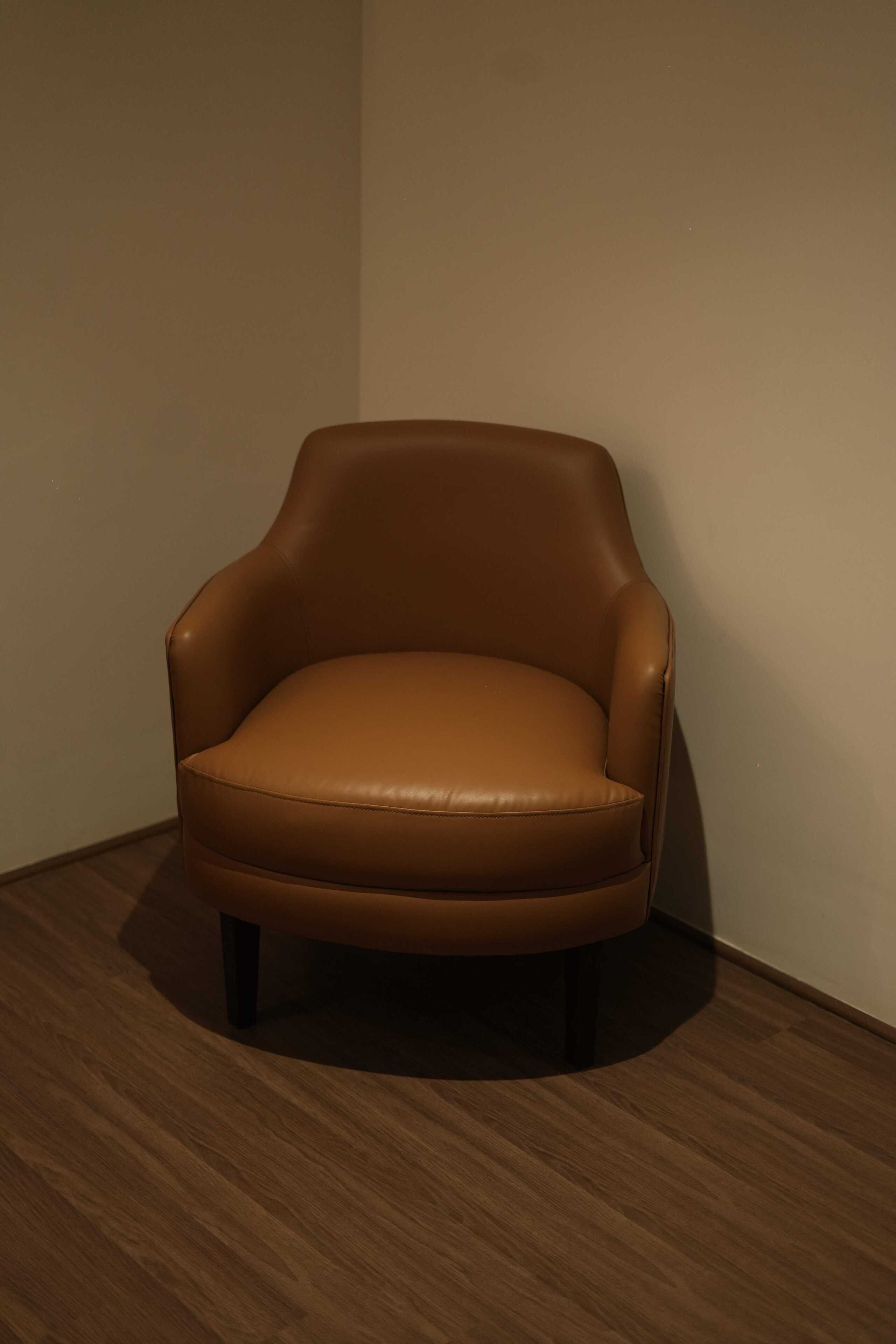 A single brown leather armchair with a high backrest and four legs, positioned against a plain wall on a wooden floor.