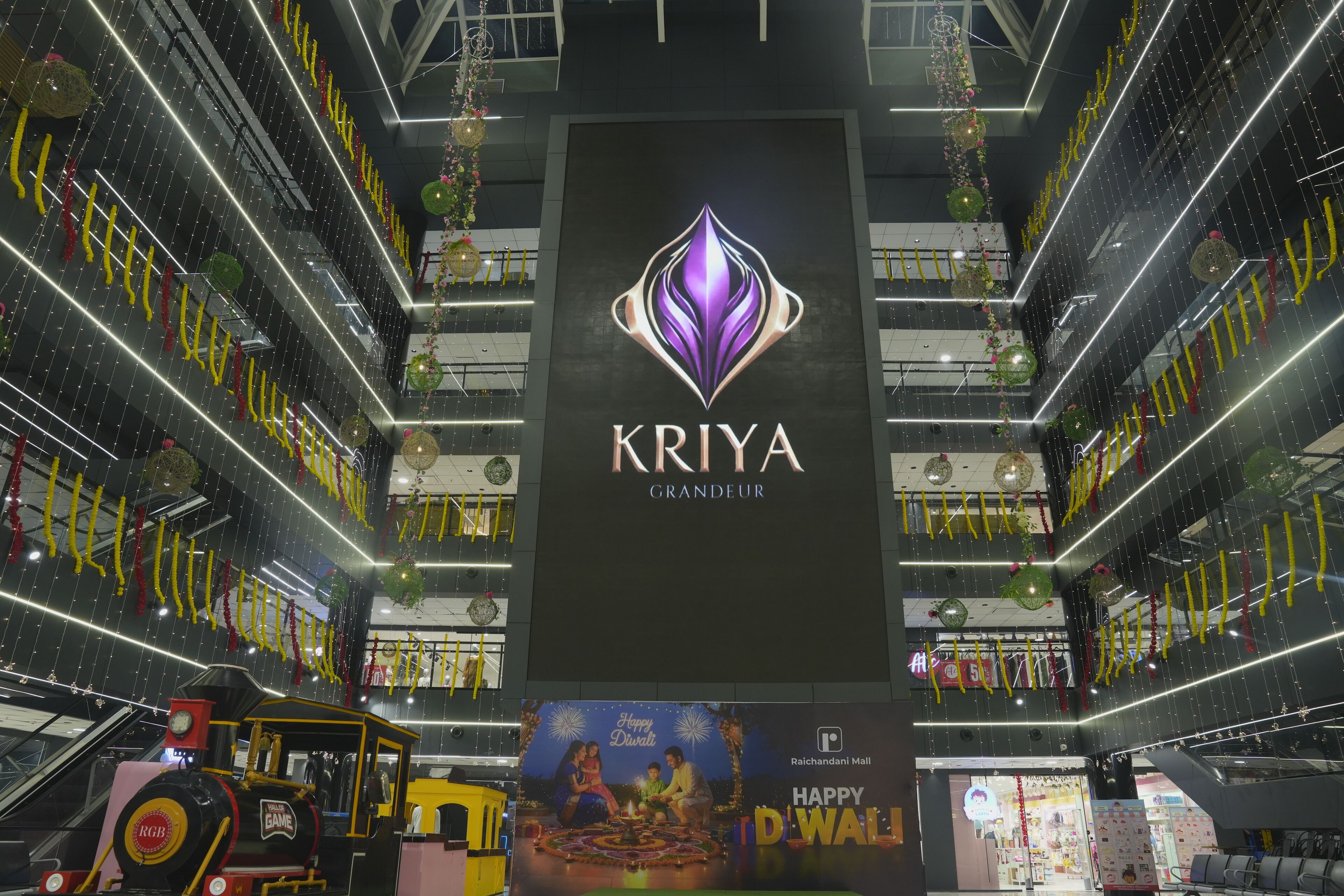 Interior de centro comercial con un llamativo letrero 'KRIIA GRANDEUR', un banderín de 'Feliz Diwali' y un paseo en tren con temática 'Feliz Diwali'.