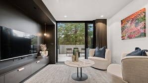 A modern living room with a flat-screen TV, a round coffee table, a sofa, and a large window offering a view of trees. - Stylish Studio in Braddon With Communal Area (Canberra)