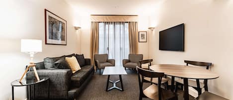 A hotel room with a sofa, two armchairs, a coffee table, a dining table with chairs, a TV, and a lamp.