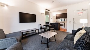 Un salon moderne avec un canapé, une table basse et un téléviseur fixé au mur. On aperçoit un coin cuisine à l’arrière-plan.