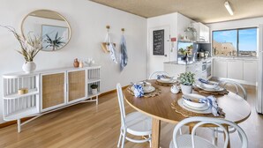 A modern kitchen with white cabinetry, a round dining table with white chairs, and a view of the city through large windows. - 2-bed Near Beach With BBQ & Stunning Ocean Views (Sunshine Coast)