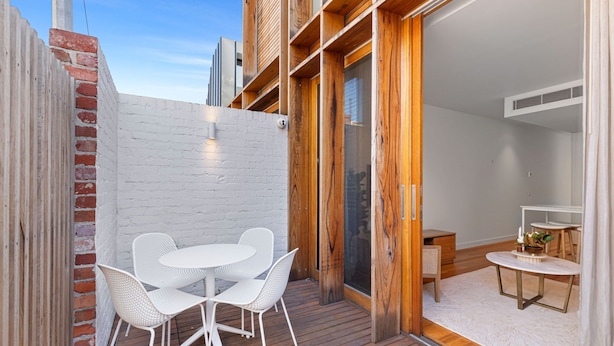 Un patio con una mesa y sillas blancas, una terraza de madera y un muro de ladrillo.