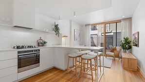 Una cocina moderna con armarios blancos, horno empotrado y estufa a gas. Hay tres taburetes de madera y una pequeña mesa de madera.
