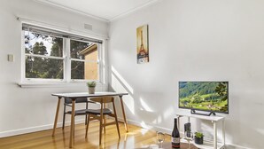 Una habitación con piso de madera, una mesa de comedor con sillas, un televisor sobre un soporte y una ventana que ofrece vistas a zonas verdes.