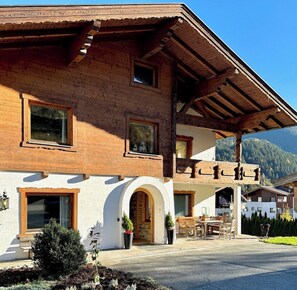 Ein zweistöckiges Holzhaus mit einem Balkon, einem steinernen Bogenportal und einem Essbereich mit Außenbestuhlung.