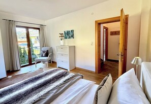 Ein Schlafzimmer mit Bett, Stuhl, Schreibtisch, einem Bild und Blick durch eine Tür.