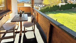 Varanda de madeira com mesa e cadeiras, vista para um gramado verde e um prédio ao fundo.