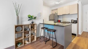 A modern kitchen with a central island, bar stools, and a wooden shelf unit with decorative items. - Luxe 2-bed Apartment With River Views Near Casino (Melbourne)