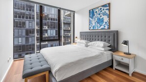 A modern bedroom with a large window, a grey upholstered bench, a neatly made bed, a bedside table with a lamp, and a floral artwork on the wall. - Luxe 2-bed Apartment With River Views Near Casino (Melbourne)