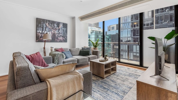 A modern living room with a grey sofa, a coffee table, and a large window offering a city view. - Luxe 2-bed Apartment With River Views Near Casino (Melbourne)