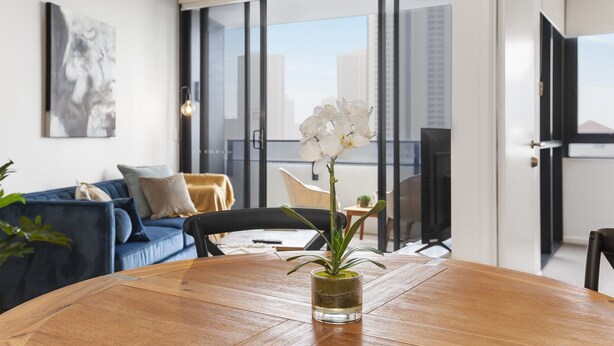 A modern living room with a wooden dining table, a glass vase with an orchid, a blue sofa, and a city view through large windows. - Ocean View 2-bed With Pool, Sauna, (Gold Coast)