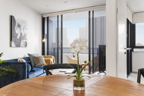 A modern living room with a wooden dining table, a glass vase with an orchid, a blue sofa, and a city view through large windows. - Ocean View 2-bed With Pool, Sauna, (Gold Coast)