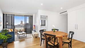 A modern living room with a wooden dining table, black chairs, a blue sofa, and a large window offering a city view. - Ocean View 2-bed With Pool, Sauna, (Gold Coast)