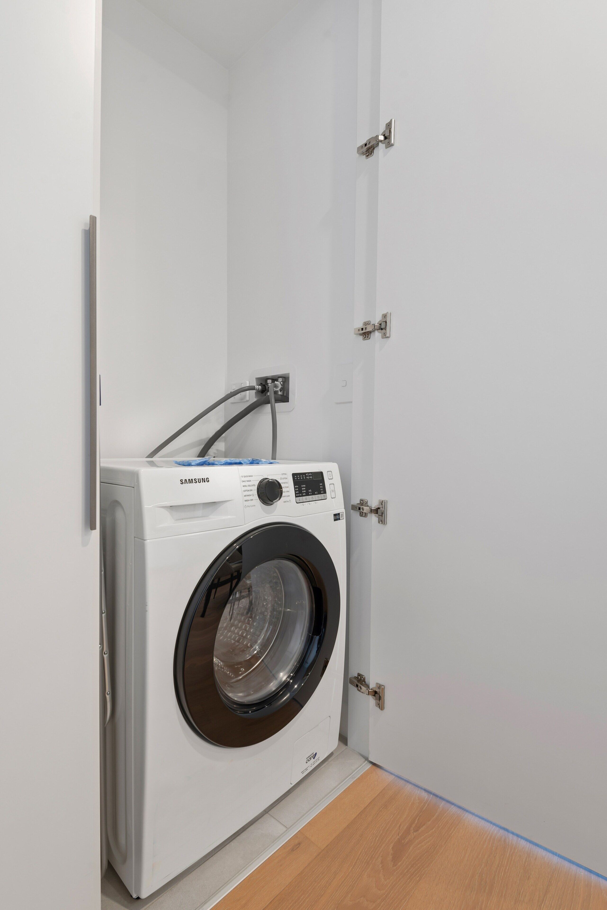 Una lavadora Samsung blanca en un lavadero pequeño y cerrado, con piso de madera y puerta blanca.