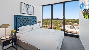Quarto com cama grande, vista da cidade e uma obra de arte emoldurada na parede.