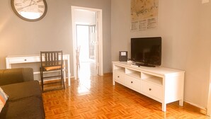 Un salon avec un plancher en bois, un téléviseur posé sur un meuble blanc, une table à manger avec des chaises et une horloge murale.