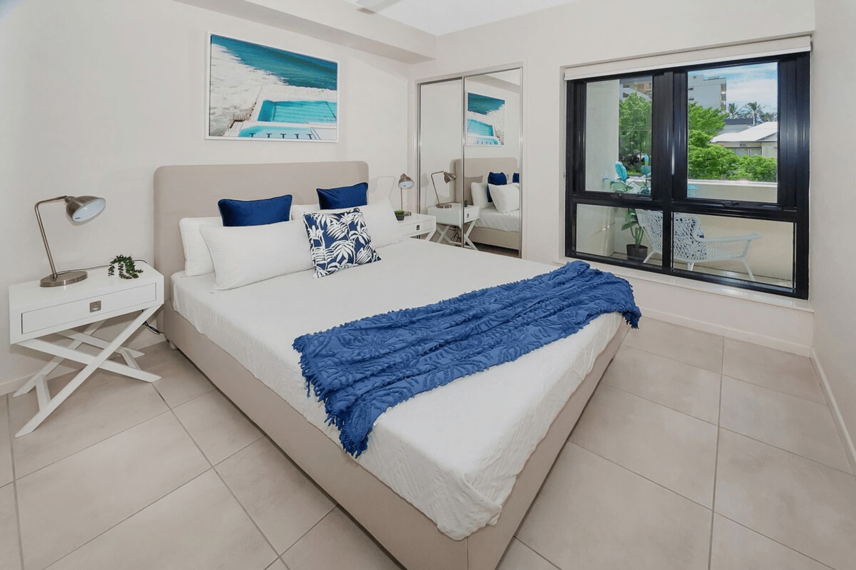 Un dormitorio con cama, mesitas de noche blancas, una manta azul, un ventanal con vistas y un cuadro en la pared.