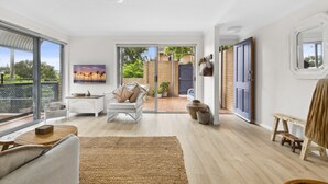 A spacious living room with a large sliding glass door leading to a backyard, a white sofa, a coffee table, and a painting on the wall. - Bright Coastal Townhouse With BBQ & Courtyard (Avoca Beach)