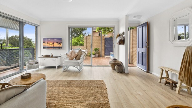 A spacious living room with a large sliding glass door leading to a backyard, a white sofa, a coffee table, and a painting on the wall. - Bright Coastal Townhouse With BBQ & Courtyard (Avoca Beach)