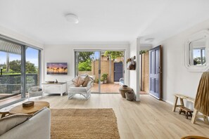 A spacious living room with a large sliding glass door leading to a backyard, a white sofa, a coffee table, and a painting on the wall. - Bright Coastal Townhouse With BBQ & Courtyard (Avoca Beach)