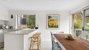 A modern kitchen with a large island, white cabinetry, and a dining area with a wooden table and blue chairs. - Bright Coastal Townhouse With BBQ & Courtyard (Avoca Beach)