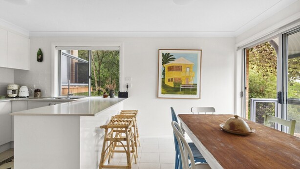 A modern kitchen with a large island, white cabinetry, and a dining area with a wooden table and blue chairs. - Bright Coastal Townhouse With BBQ & Courtyard (Avoca Beach)