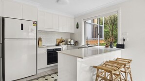 A modern kitchen with white cabinetry, a built-in island, and a large window offering a view of the outdoors. - Bright Coastal Townhouse With BBQ & Courtyard (Avoca Beach)