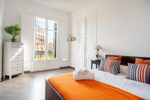Quarto de hotel com cama, penteadeira, mesa de cabeceira, abajur e vista para edifícios pela janela.